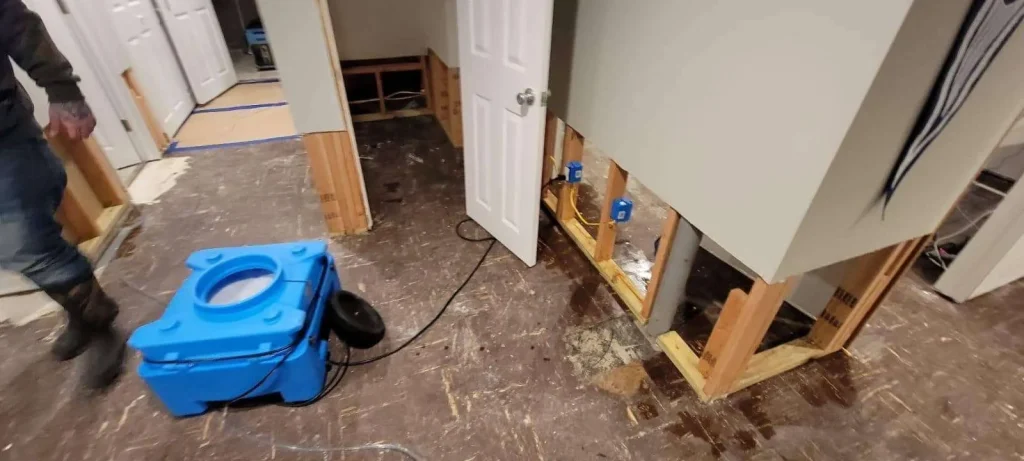 A room with damaged flooring and cut-out drywall, exposing wall studs and electrical boxes, shows clear signs of water damage. A blue drying machine is on the floor, and a person in boots stands to the left.